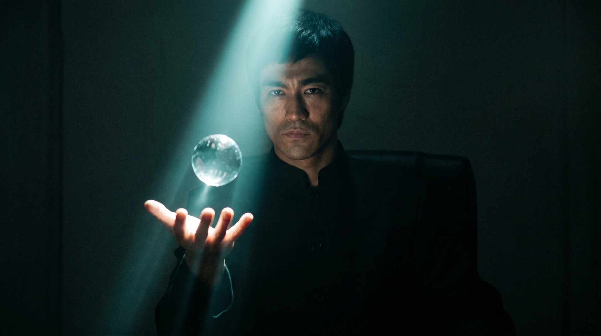 A man cradling a clear glass orb in his outstretched palm, lit by a single cool beam of light slicing through near-black darkness — the brand DNA as water held in the model's vessel.